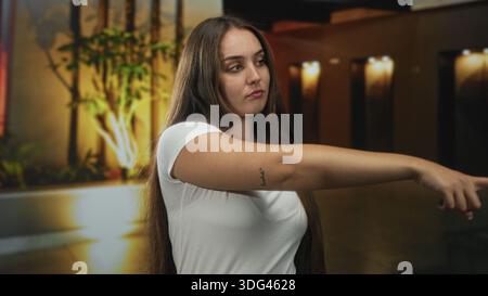 Frau posiert mit sichtbarem Dekolleté und langen Haaren, das Kinn im Flur der Hotellobby durch beleuchtete Pflanzgefäße und Flurlichter erhöht; Selbstvertrauen. Stockfoto
