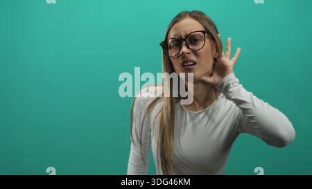 Frau mit Brille, die Hand an Ohr schröpft und den Kopf leicht dreht im grünen Studio; Neugier Hörfokus Konzentration. Stockfoto
