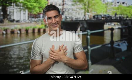 Mann mit Händen, die auf einer amsterdamer Straße an einem Kanal an der Brust gefesselt sind, sanft lächelnd; Dankbarkeit verbunden. Stockfoto