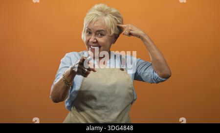 Die ranghöchste hispanische Frau in der Schürze zeigt den Finger auf die Kamera und zeigt den Finger zum Tempel in einem Studio mit orangefarbenem Hintergrund; spielerisches Selbstvertrauen. Stockfoto