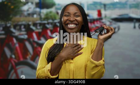 Die lächelnde Frau in einem hellgelben Hemd hält Autoschlüssel, die draußen neben Mietfahrrädern auf einer Stadtstraße mit verschwommenem Hintergrund stehen. Stockfoto