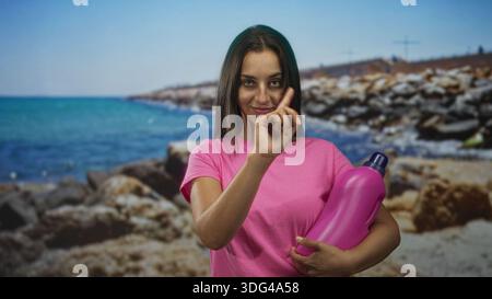 Junge Frau, die mit dem Zeigefinger auf die felsige Küste zeigt und eine große pinkfarbene Waschmittelflasche hält; verspielte Putzbotschaft. Stockfoto