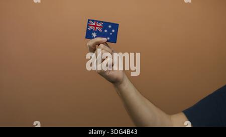 Ein Mann hält eine kleine australische Flagge an der braunen Wand mit Fokus auf Stickdetails und zeigt Nationalstolz und Identität in einem einfachen, isolierten Set Stockfoto