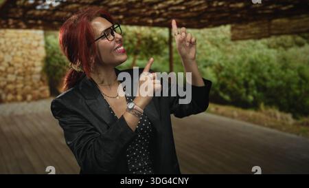 Frau zeigt den Zeigefinger nach oben auf die Veranda, lächelt mit Brillen und mehrschichtigen Ketten; Freude am Vertrauen. Stockfoto