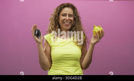 hispanische Frau mittleren Alters, die Autoschlüssel in der linken Hand und gelbe Schweinebank in der rechten Hand hält, während sie im Studio lächelt; Vertrauen in die finanzielle Planung. Stockfoto