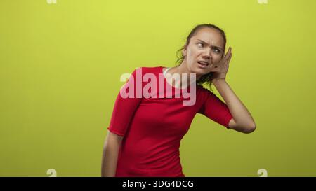 Frau, die mit der Hand das Ohr schröpft, den Kopf neigt, sich vor die lindgrüne Studiowand lenkt und dabei rotes Hemd trägt; Neugier-Aufmerksamkeit. Stockfoto