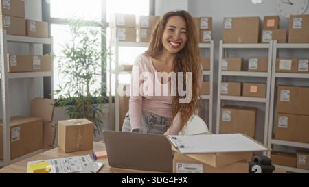 Frau, die auf einem Laptop schreibt, während sie Paketetiketten an einem Schreibtisch im Gebäude überprüft; Produktivität kleiner Unternehmen. Stockfoto