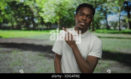 Mann mit weißem Polohemd und Brille von Hand zu Ohr, der im Wald unter sonnendurchflutetem Baldachin Telefongeste macht; Kommunikationsfreude. Stockfoto