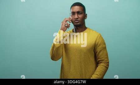 Junger schwarzer Mann mit Brille und gelbem Pullover, Smartphone an Ohr hält und im Studio mit der rechten Hand in Frage stellt; Verwirrung. Stockfoto