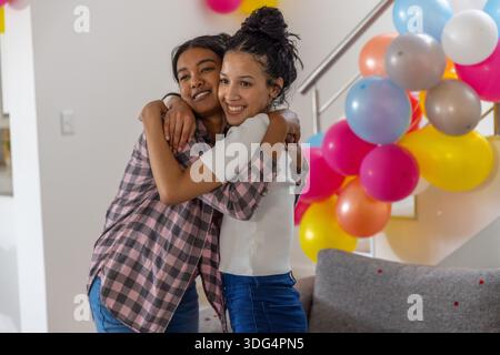 Diverse Mutter und Teenager-Tochter umarmen sich in der Nähe der Treppe im Wohnzimmer mit Ballons Stockfoto