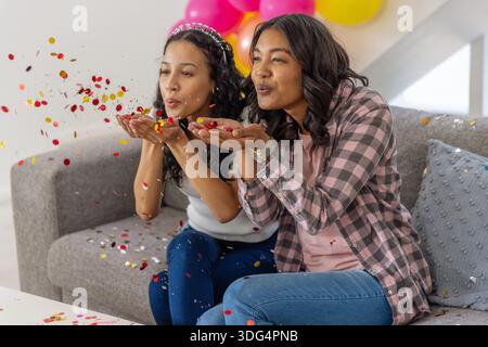 Diverse Mutter und Teenager-Tochter blasen rot, Gold und Konfetti im Wohnzimmer mit Ballons Stockfoto