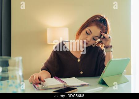 Junge Frau, die geistige Ermüdung erleidet, während sie drinnen an einem Tablet und Notebook arbeitet und Stress, Konzentration, kognitive Überlastung und Druck zeigt Stockfoto