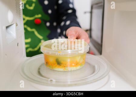 Person in einem Weihnachtspullover, die einen Plastikbehälter mit Gemüsesuppe in eine Mikrowelle stellt Stockfoto