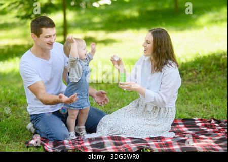 Ein Kleinkind streckt nach Seifenblasen, die seine Mutter bläst. Die Familie genießt einen sonnigen Tag mit verspieltem Baby und Seifenblasen, bei einem Picknick im Freien Stockfoto
