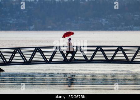 Eine Person mit einem roten Schirm geht über eine Metallbrücke über ruhiges Wasser. Bainbridge Island, WA, USA Stockfoto