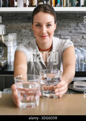 Frau in einem Poloshirt, die Gläser Wasser mit einem Lächeln in einer modernen Küche anbietet. Deutschland Stockfoto