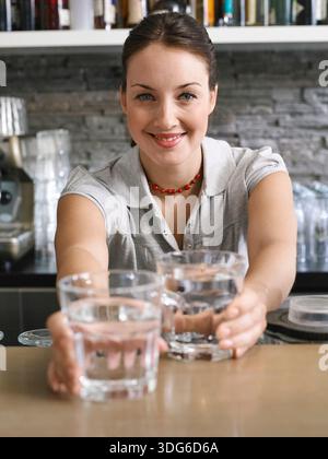 Lächelnde Frau in grauem Hemd, die drinnen zwei Gläser Wasser über eine Theke anbietet. Deutschland Stockfoto