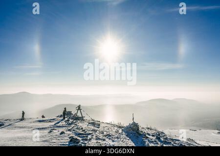 Sonnenhunde über verschneiten Bergketten mit Menschen, die die Winterlandschaft bewundern. Reiseerlebnis bei kaltem Wetter für Erkundungskonzept. Stockfoto
