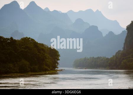 Ruhige Flusslandschaft, umgeben von üppigen Bäumen und fernen Bergsilhouetten unter einem weichen Himmel. Li River, Li Jiang, Guangxi, China Stockfoto
