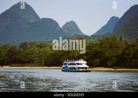 Ein Boot segelt auf einem Fluss mit üppigen grünen Hügeln und nebeligen Bergen im Hintergrund. Li River, Li Jiang, Guangxi, China Stockfoto
