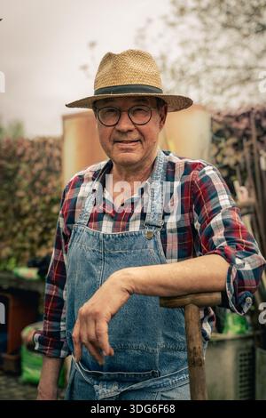 Seniorgärtner mit Denim-Schürze im Freien in einem Gemüsegarten, Porträt eines aktiven älteren Landwirts und nachhaltiger ländlicher Lebensstil. Stockfoto