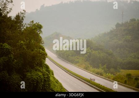Eine ruhige, leere Straße, umgeben von üppigen grünen Hügeln und Bäumen an einem nebeligen Tag. Ausflug von Guilin nach Chengdu, Guangxi, Sichuan, China Stockfoto