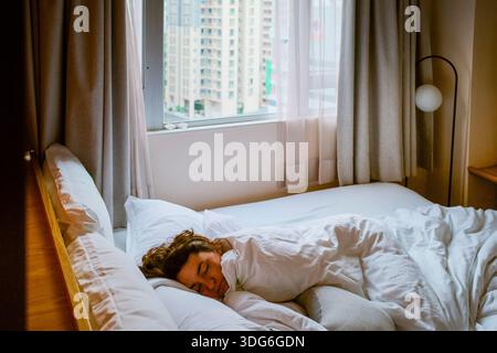 Eine Person, die friedlich in einem hellen, gemütlichen Schlafzimmer mit Blick auf die Stadt durch das Fenster schläft. Hongkong, China Stockfoto