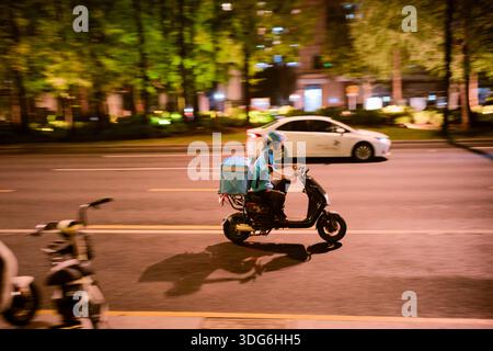 Der Lieferer fährt mit einem Roller nachts auf einer Stadtstraße mit leuchtenden Lichtern. Shanghai, China Stockfoto
