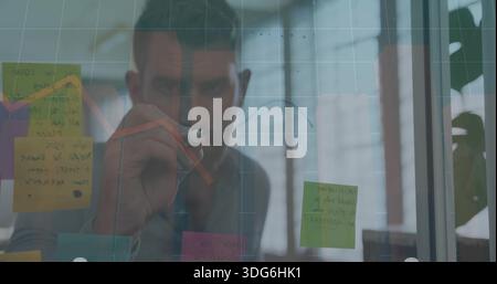 Ein Mann in grauem Hemd, der eine Glasplatte im Büro beschriftet, einen Marker hält, Haftnotizen, einen roten Pfeil Stockfoto