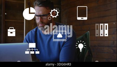 Arbeitender bärtiger Mann mit Brille, blauem Pullover am Laptop im Holzzimmer mit grüner Jacke, Ikonen Stockfoto