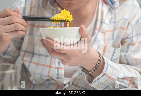 Mann, der eine Schüssel hält und mit Essstäbchen frühstückt. Junger Mann, der Essstäbchen hält und traditionelles asiatisches Essen isst. Nahaufnahme, selektiver Fokus Stockfoto