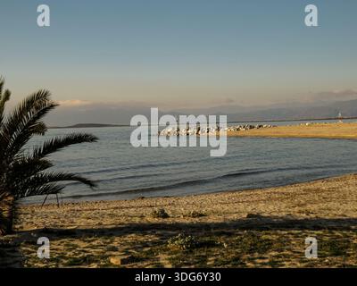 Ruhige Küstenlandschaft mit Sandstrand, ruhigem Meer und nebligen Bergen unter einem klaren blauen Himmel am frühen Morgen. Stockfoto