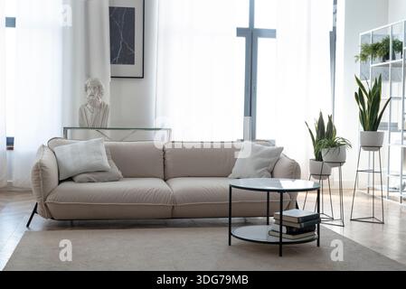 Modernes, helles Wohnzimmer mit beigefarbenem Sofa, Marmor-Couchtisch, grünen Pflanzen und einer klassischen Büstenstatue Stockfoto