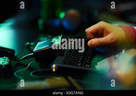 Kaukasischer junger erwachsener Mann, der USB-Flash-Laufwerk in die Computertastatur auf einem dunklen Schreibtisch einsteckt, von Hand sichtbar, um Hacker-Aktivitäten auszuführen, elektronische Geräte und Kabel um den Arbeitsplatz herum Stockfoto