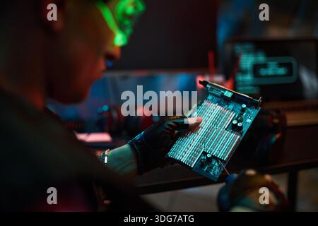 Junger Mann mit Handschuhen, der Computerplatine hält und Hardware mit verschwommenem Laptop überprüft, der Code im Hintergrund anzeigt, der auf Cybersicherheit oder Hackeraktivitäten hindeutet Stockfoto