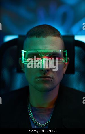 Porträt eines jungen Erwachsenen kaukasischen Mannes mit futuristischem Neonvisier, der in einem dunklen Raum sitzt, Gesichtspiercings sichtbar, direkt in die Kamera blickend, beleuchtet durch grüne und blaue Lichter Stockfoto