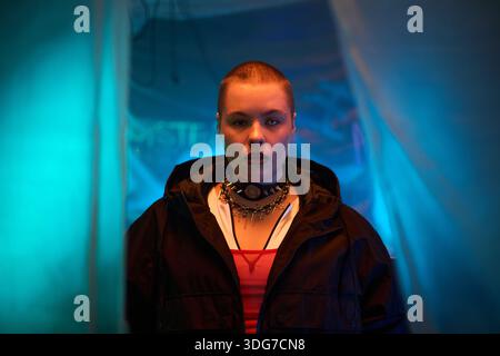Porträt einer jungen kaukasischen Frau mit rasiertem Kopf und Gesichtspiercings, die in schwach beleuchtetem Raum steht, mit einem Halsband und einer dunklen Jacke, die direkt in die Kamera blickt Stockfoto