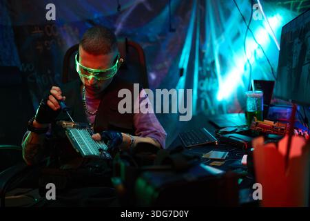 Junger Erwachsener kaukasischer Mann mit futuristischer Brille, am Schreibtisch sitzt, Computersystem hackt und Hardware modifiziert, mit Schraubenzieher und Tastatur, umgeben von elektronischen Geräten und Neonlichtern Stockfoto