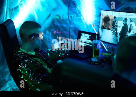 Junger Erwachsener kaukasischer Mann mit futuristischer Brille, der am Schreibtisch sitzt und ein Computersystem hackt, beleuchtet durch blaue Neonlichter, mehrere Monitore, die Code und Überwachungsaufnahmen anzeigen Stockfoto