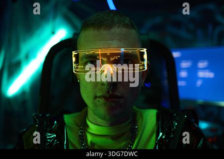 Porträt eines jungen Erwachsenen kaukasischen Mannes mit futuristischem Cyberpunk-Neonvisier, der in einem dunklen Raum mit leuchtenden Bildschirmen sitzt, Gesichtspiercings sichtbar, direkt in die Kamera blickt, Hackerkonzept Stockfoto