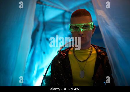 Porträt eines jungen Erwachsenen kaukasischen Mannes mit futuristischer Neonbrille, der in schwach beleuchtetem Raum mit blauer Beleuchtung steht, direkt in die Kamera blickt und das Konzept des Cyberpunk-Hackers verkörpert Stockfoto