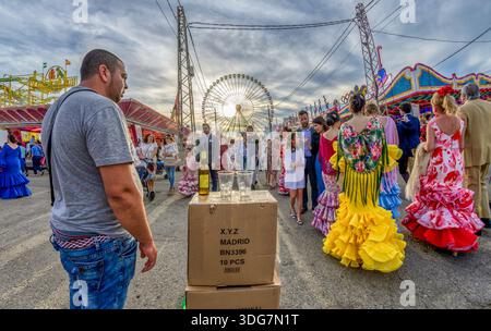 Straßenverkäufer mit Getränken zum Verkauf auf der Aprilmesse in Sevilla. Personen gehen in traditioneller Flamenco-Kleidung und moderner Kleidung am Verkäufer vorbei. Stockfoto