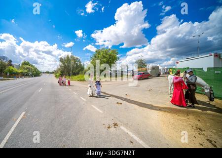 Reiter und Frauen in Flamenco-Kleidern laufen von den umliegenden Straßen aus unter teilweise bewölktem Himmel an einem sonnigen Tag zur Feria de Abril in Sevilla. Stockfoto
