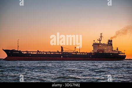 Ein Frachtschiff fährt während eines lebhaften Sonnenuntergangs durch die Industriezone der Flussmündung des Odiel in Huelva, Spanien. Dramatische maritime Szene. Stockfoto