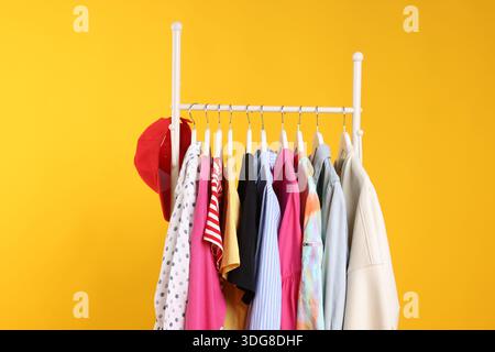 Verschiedene Kleidung und Baseballmütze hängen auf dem Regal vor orangem Hintergrund, Nahaufnahme Stockfoto