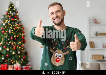 Junger Mann im Weihnachtspullover, der zu Hause die Daumen nach oben zeigt Stockfoto