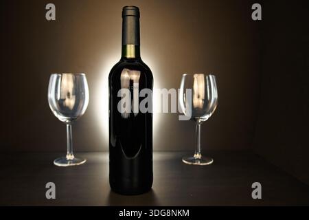 Hintergrundbeleuchtete Weinflasche und zwei leere Gläser auf dunklem Hintergrund, elegantes Studiostilleben mit Kopierraum. Stockfoto