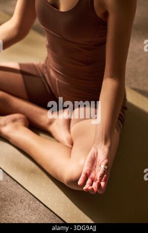 Junge Erwachsene Frau, die im Kreuz auf einer Yogamatte sitzt und mit geschlossenen Augen meditiert, die Hand in entspannter Position auf dem Knie ruht, Achtsamkeit in Innenräumen übt Stockfoto