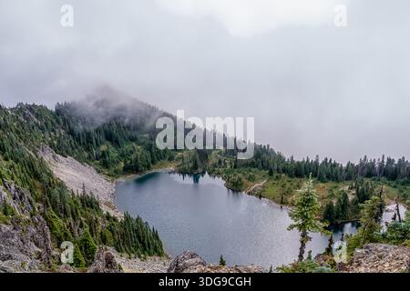 Ein großes Gewässer, umgeben von einem üppigen Wald und hohen Bergen. Der Himmel darüber ist bewölkt und schafft eine stimmungsvolle Atmosphäre. Die Szene ist heiter und Stockfoto