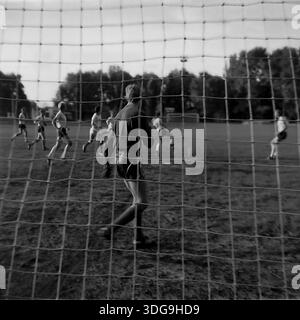 Slawjansk, Region Donezk, Ukraine, UdSSR. Archiv Schwarzweiß-Foto aus den 1980er Jahren, das ein Fußballspiel im Khimik Stadium durch das Tornetz zeigt. Das Bild zeigt einen Torhüter im Vordergrund, der das Spiel auf dem Rasenfeld aufmerksam beobachtet. Andere Spieler sind in der Distanz sichtbar, laufen und konkurrieren. Das Netznetz rahmt die Szene ein und verleiht der Komposition zusätzliche Tiefe. Hohe Parkbäume umgeben den Sportplatz. Diese Momentaufnahme veranschaulicht die Beliebtheit des Fußballs und der aktiven Freizeit in der friedlichen Industriestadt vor dem Konflikt. Stockfoto